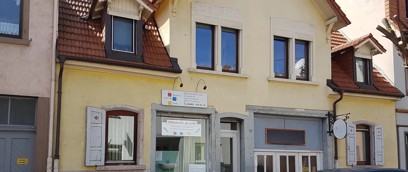 Die Ladenfront an der Friedrichstraße von der Straße aus: gelbe Fassade, rotes Ziegeldach, helle Klappläden und das Firmenschild über dem Schaufenster.