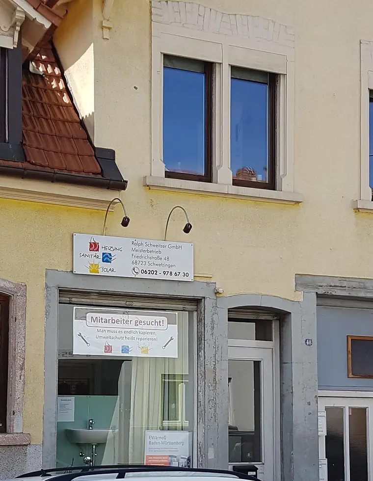 Das Haus in der Friedrichstraße 48 im Ausschnitt: Ziegeldach, zwei Fenster, darunter das Firmenschild und das Schaufenster mit dem Aushang.