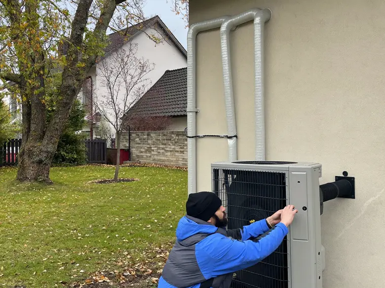 Außeneinheit einer Luft-Wasser-Wärmepumpe auf zwei Betonsockeln an der Hauswand, daneben ein Monteur, der eine Verkleidungsschraube setzt.