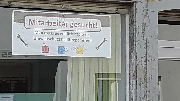 Der Aushang im Schaufenster: eine Stellenanzeige, darunter der Satz über das Reparieren, links und rechts ein gemalter Schraubenschlüssel.