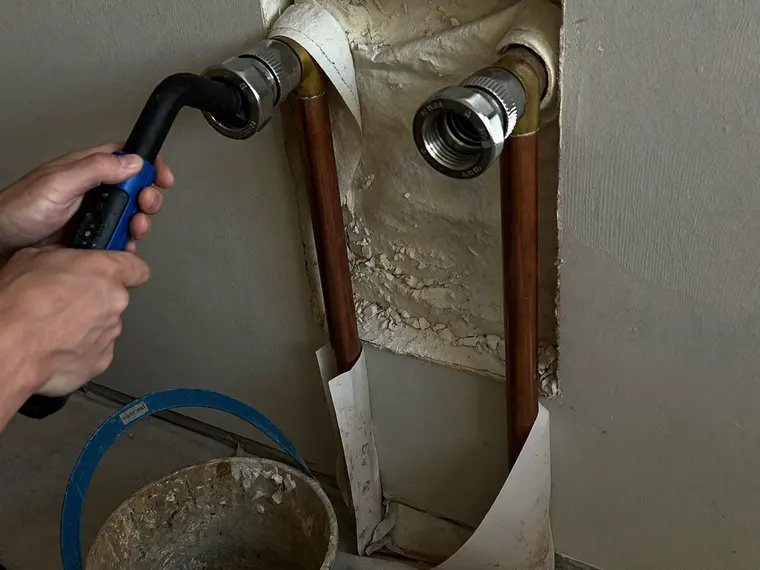 Geöffnete Wandöffnung mit zwei Kupferleitungen und frisch verpressten Fittings, davor ein Presswerkzeug in den Händen eines Installateurs.