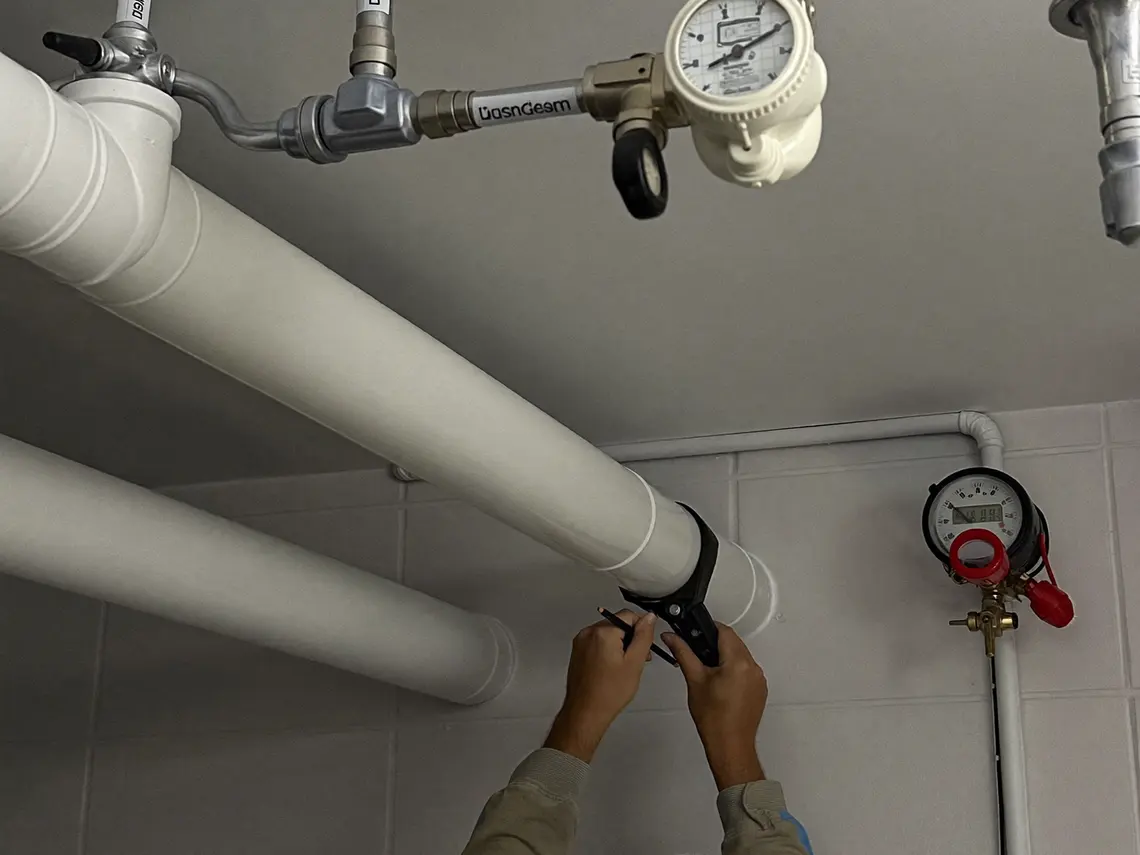 Neue gedämmte Wasserleitungen unter einer Kellerdecke mit beschrifteten Verteilern, Absperrventilen und Wasserzähler, darunter ein Installateur beim Anzeichnen.